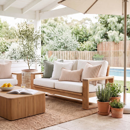 Outdoor patio furniture set including a wooden loveseat with cream cushions and striped pillows, a matching wooden coffee table, and a side chair under a patio umbrella with a garden background.