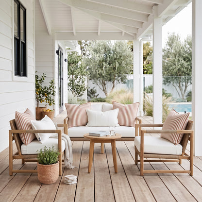 A three-piece outdoor patio furniture set with light wood frames, white cushions, and striped pink and beige pillows, arranged on a wooden porch with a small round wooden coffee table.
