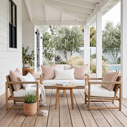 A three-piece outdoor patio furniture set with light wood frames, white cushions, and striped pink and beige pillows, arranged on a wooden porch with a small round wooden coffee table.