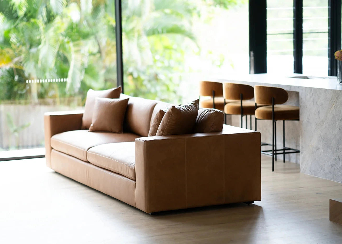 Brown leather sofa in a modern living room with large windows and a kitchen area.