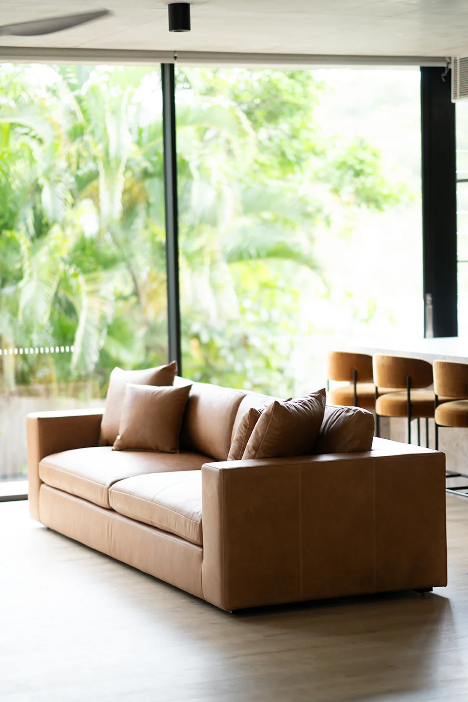 Brown leather sofa with cushions in a room with large windows showing greenery outside.