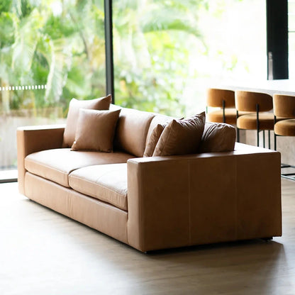 Brown leather sofa with cushions in a room with large windows showing greenery outside.
