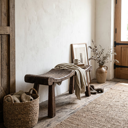 A rustic wooden bench sits in a sunlit room corner against a textured white wall, with a beige throw draped over it, a woven basket, shoes, a ceramic vase with dried branches, and a framed print nearby.