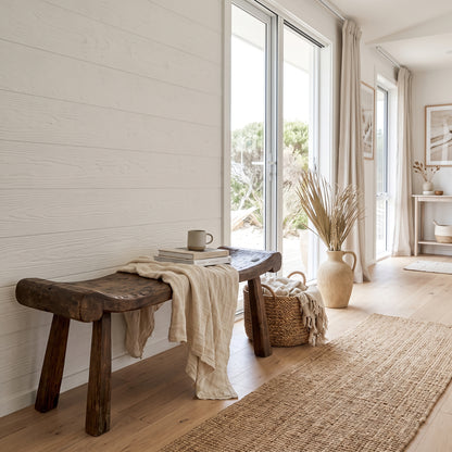 A rustic dark wood bench in a bright, modern coastal-style room with white shiplap walls, a beige linen throw, a woven basket, and a large vase of dried grasses near tall glass doors.