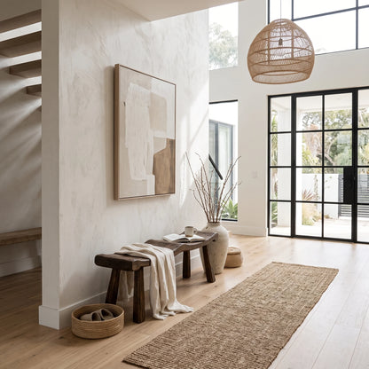 A modern, sunlit entryway with a large abstract art print on a textured white wall, a dark wooden bench with a draped throw, a woven jute runner, and a large round wicker pendant light hanging from the ceiling.