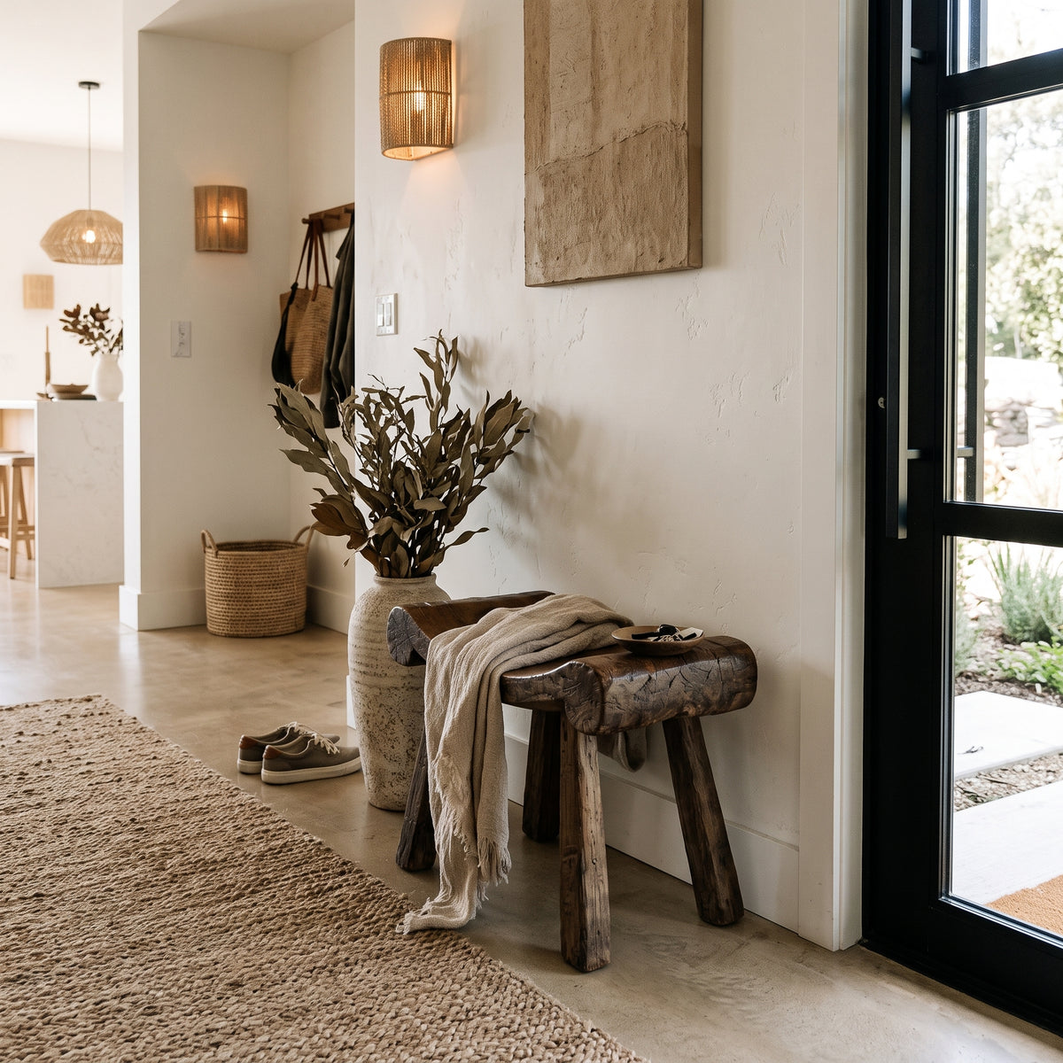 A rustic wooden bench with three legs sits in a bright entryway next to a large vase of dried leaves, with a beige throw blanket draped over it. The scene features a textured rug, a black-framed glass door, and warm wall lighting.