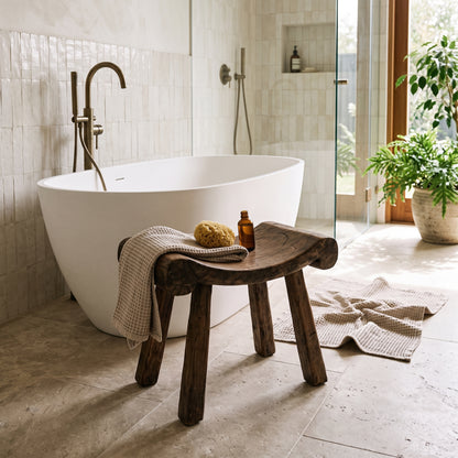 A freestanding oval bathtub in a bright, modern bathroom with light stone tiles, a dark wooden stool holding a folded towel and small amber bottle, and a glass shower enclosure in the background.