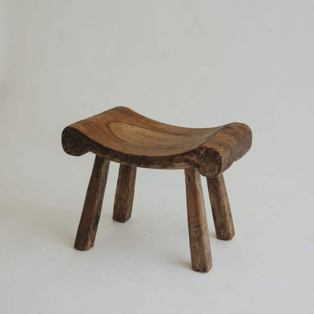 A rustic, hand-carved wooden stool with a curved seat and three legs, shown on a plain light gray background.