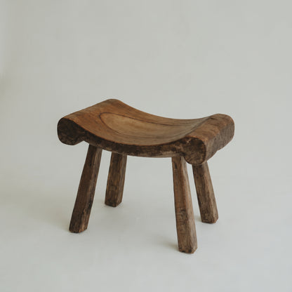 A rustic, hand-carved wooden stool with a curved seat and three legs, shown on a plain light gray background.