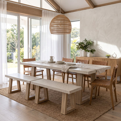 A modern dining set in a bright, sunlit room, featuring a rectangular light stone-top table with a thick wooden base, two matching stone-and-wood benches, and four woven-back wooden chairs. A large wicker pendant light hangs above, and a sideboard with plants is in the background.