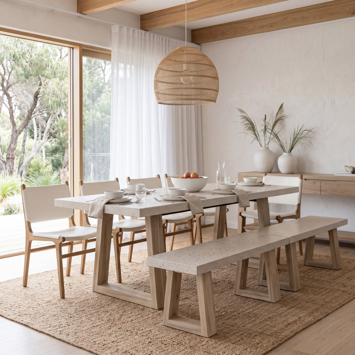 A modern dining set in a bright, sunlit room, featuring a rectangular light stone-top table with a thick wooden base, two matching stone-and-wood benches, and four woven-back wooden chairs. A large wicker pendant light hangs above, and a sideboard with plants is in the background.