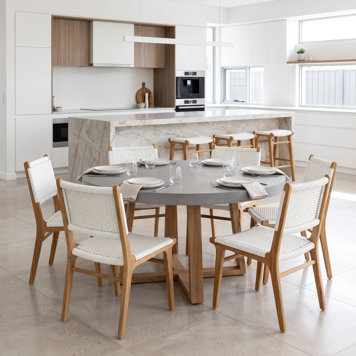 A round gray dining table with six wooden chairs featuring woven off-white seats, set in a bright, modern kitchen with a marble island and light wood cabinetry.