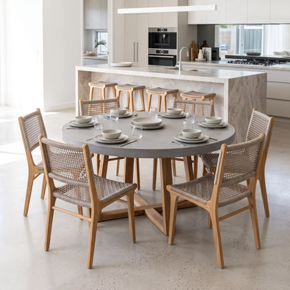 A round dining table with a gray stone-look top and light wood frame, surrounded by six matching woven-back chairs in a bright modern kitchen setting.