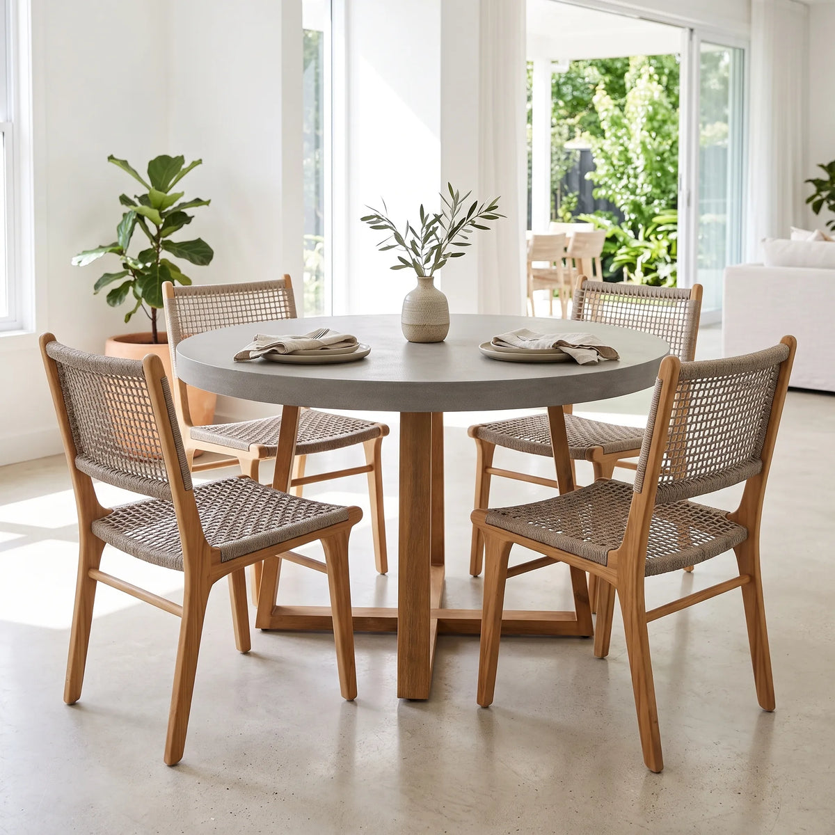 A round dining table with a light gray stone top and four wooden chairs with woven cane seats, set in a bright, modern dining room with large windows and plants.
