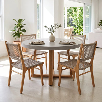 A round dining table with a light gray stone top and four wooden chairs with woven cane seats, set in a bright, modern dining room with large windows and plants.