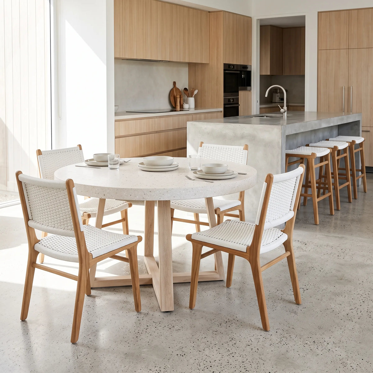 A round dining table with a light stone top and natural wood legs, surrounded by four woven-back wooden chairs with white seats, set in a bright modern kitchen with wood cabinetry and a concrete island.