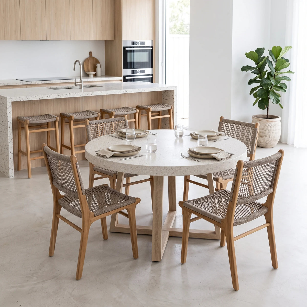 A round dining table set with six woven-back wooden chairs in a bright, modern kitchen and dining room. The table has a light beige stone top and a light wood pedestal base, while the chairs feature light oak wood frames and woven cane seats.