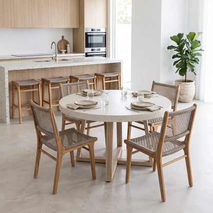 A round dining table set with six woven-back wooden chairs in a bright, modern kitchen and dining room. The table has a light beige stone top and a light wood pedestal base, while the chairs feature light oak wood frames and woven cane seats.