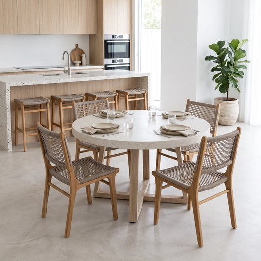 A round dining table set with six woven-back wooden chairs in a bright, modern kitchen and dining room. The table has a light beige stone top and a light wood pedestal base, while the chairs feature light oak wood frames and woven cane seats.