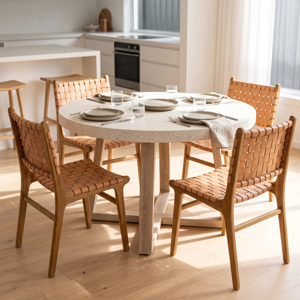 A round dining table with a light beige stone-look top and natural wood legs, surrounded by four woven-back chairs in a bright, modern kitchen setting.