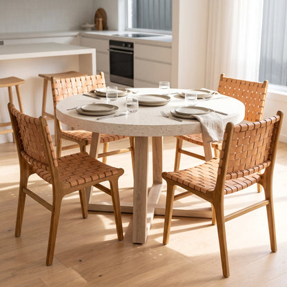 A round dining table with a light beige stone-look top and natural wood legs, surrounded by four woven-back chairs in a bright, modern kitchen setting.