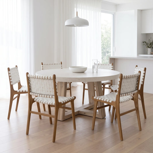 A round white dining table with a light wood pedestal base, surrounded by six wooden chairs with woven cream and white seat and back panels, set in a bright, modern white dining room.