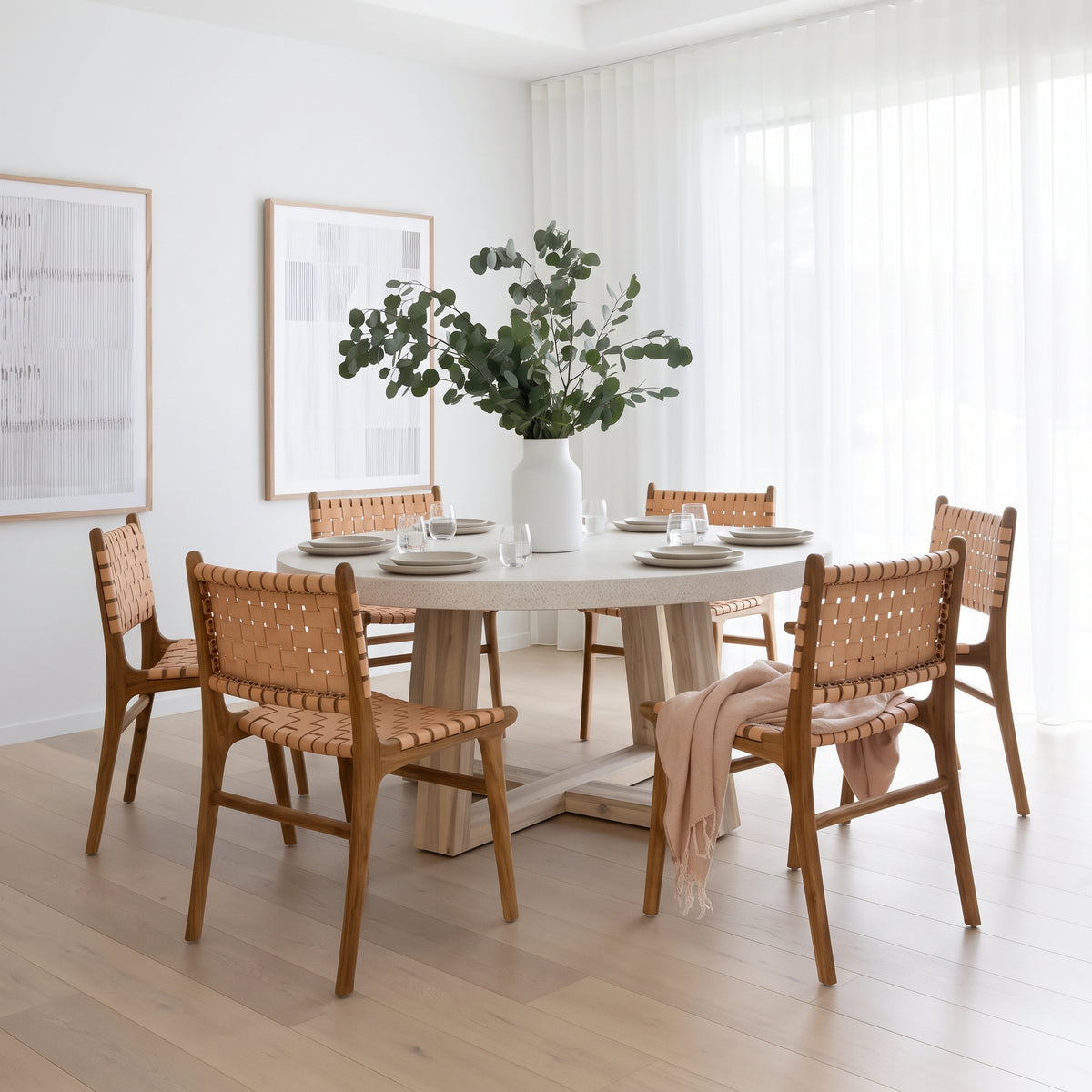 A round dining table with a light stone top and thick wooden pedestal base, surrounded by six woven-back chairs in a bright, modern dining room. A large white vase filled with green foliage sits on the table, which is set with white plates and glasses.