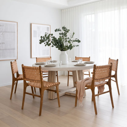 A round dining table with a light stone top and thick wooden pedestal base, surrounded by six woven-back chairs in a bright, modern dining room. A large white vase filled with green foliage sits on the table, which is set with white plates and glasses.