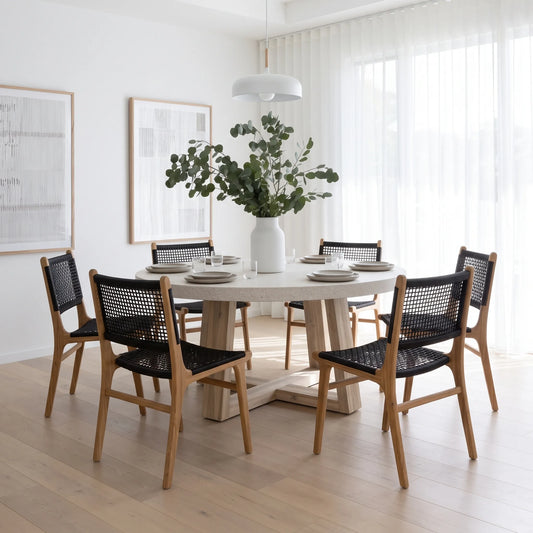 A round dining table with a light wood base and six matching chairs featuring black woven seats and backs, set in a bright, modern dining room with white walls, sheer curtains, and a large white vase of greenery in the center.