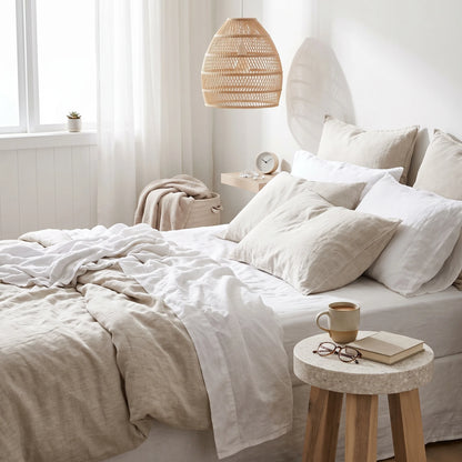 A neatly made bed with white and beige linen bedding in a bright, minimalist bedroom. A woven pendant light hangs above, and a small wooden side table holds a mug, book, and glasses.