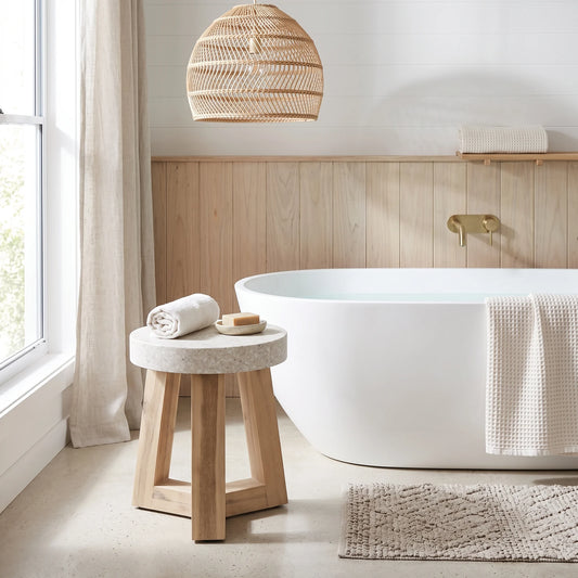 A modern bathroom scene featuring a white freestanding bathtub, a small round stone-and-wood stool with a rolled towel, a woven wicker pendant light, light wood wainscoting, and a neutral bath mat.