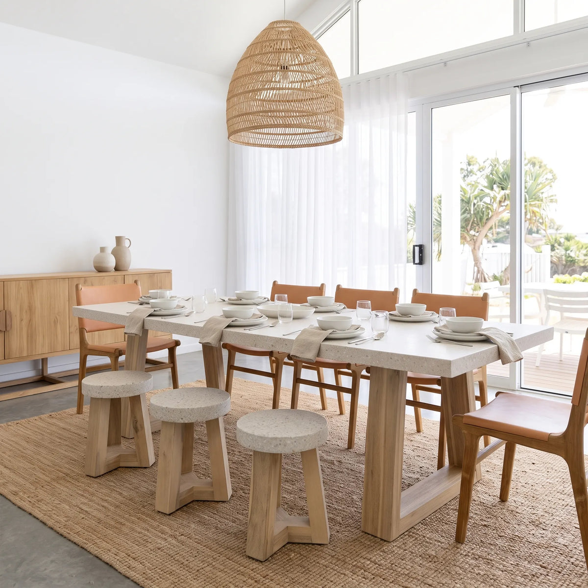 A modern dining room setup featuring a rectangular light wood table with a white stone top, surrounded by wooden chairs and three matching stone stools, set on a large woven jute rug.