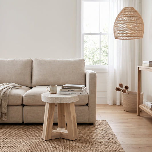 A light beige fabric sofa in a bright, modern living room setting next to a small round wooden side table with a stone top.