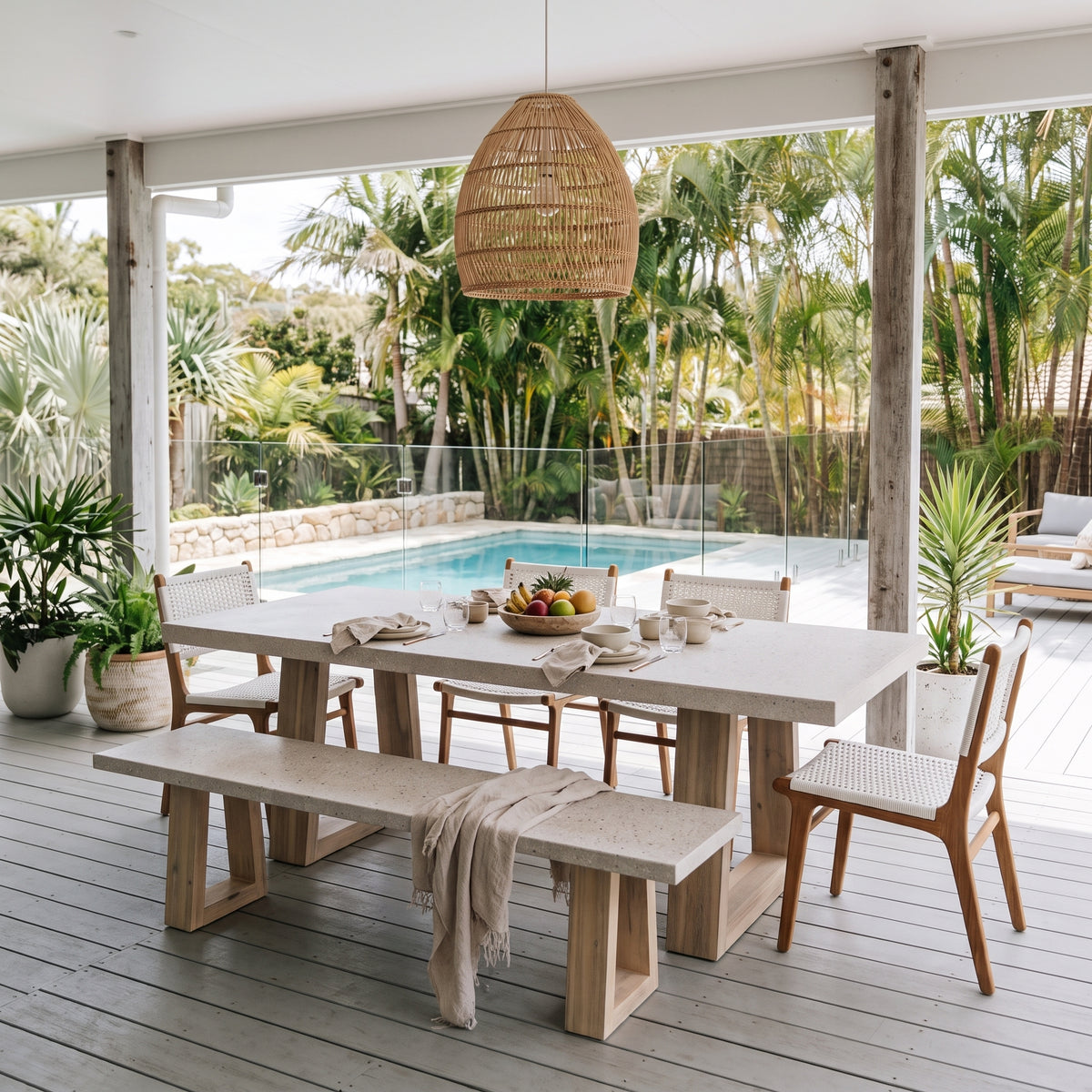 A modern outdoor dining set featuring a rectangular light stone table, a matching bench, and woven-back chairs, arranged on a gray deck under a wicker pendant light with a swimming pool and tropical garden in the background.