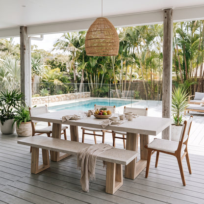 A modern outdoor dining set featuring a rectangular light stone table, a matching bench, and woven-back chairs, arranged on a gray deck under a wicker pendant light with a swimming pool and tropical garden in the background.
