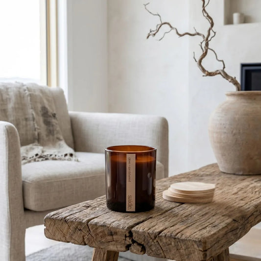 Brown candle on a wooden table with a neutral-colored sofa and decorative vase in the background.