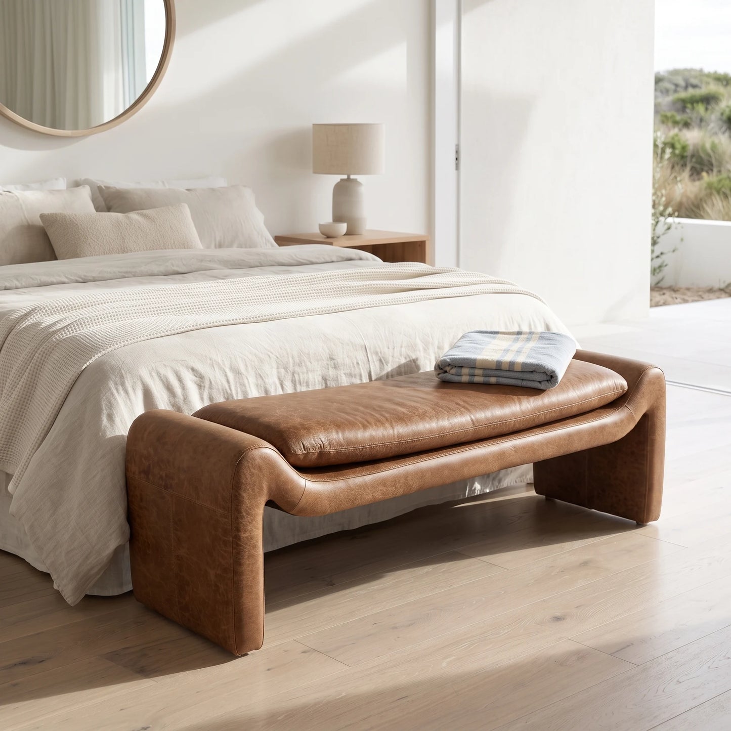 A brown leather bench with curved arms and a padded seat cushion sits at the foot of a neatly made bed in a bright, modern bedroom.