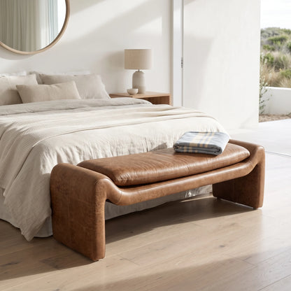 A brown leather bench with curved arms and a padded seat cushion sits at the foot of a neatly made bed in a bright, modern bedroom.
