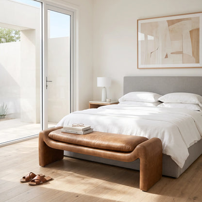 A modern bedroom scene featuring a brown leather bench with curved arms at the foot of a bed with white linens and a gray upholstered headboard. Large glass sliding doors are visible on the left, and a pair of brown leather sandals sits on the wooden floor nearby.
