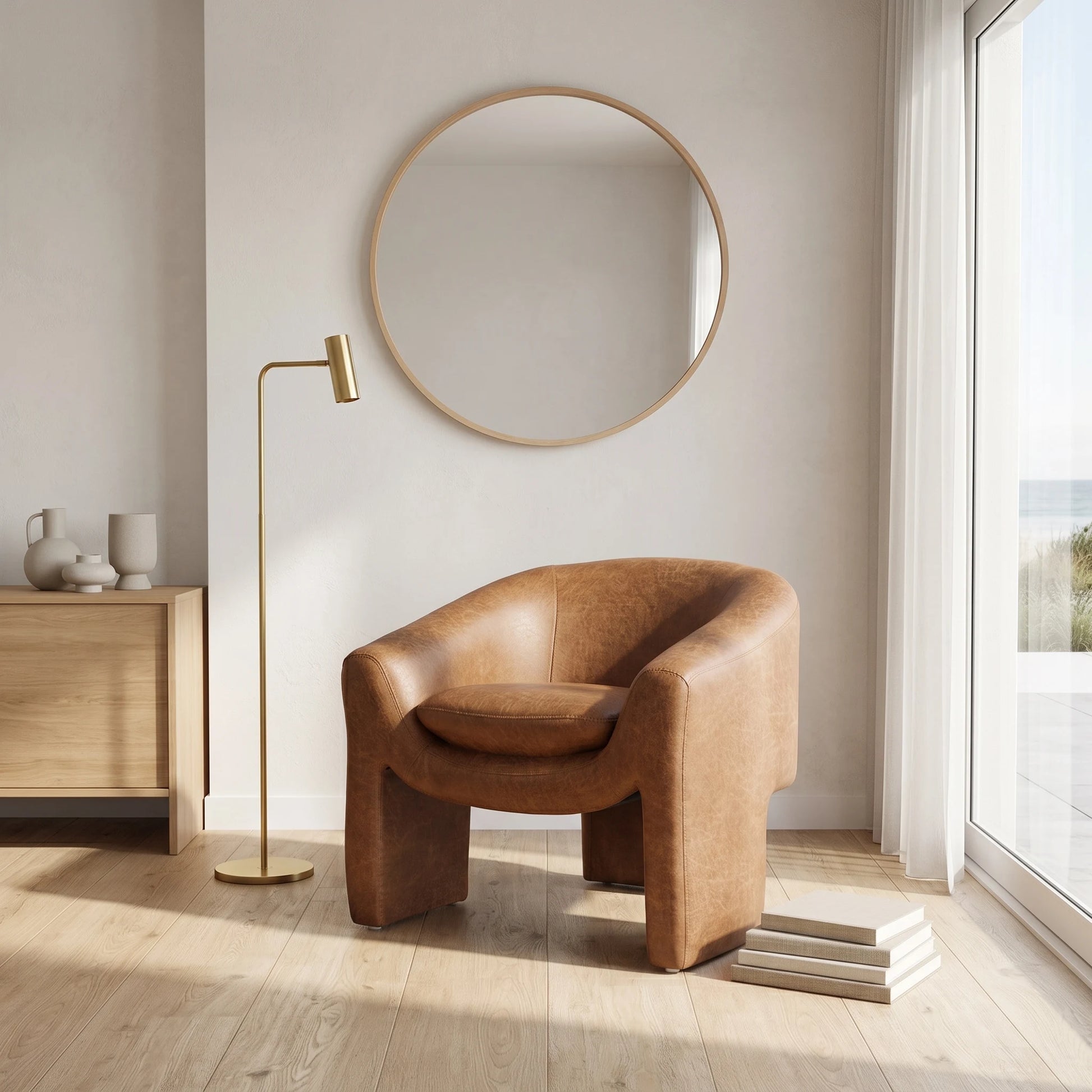 A modern tan leather barrel chair in a bright, minimalist room with a gold floor lamp, a round wood-framed mirror, and light wood flooring.