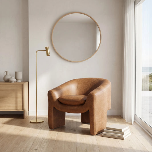 A modern tan leather barrel chair in a bright, minimalist room with a gold floor lamp, a round wood-framed mirror, and light wood flooring.