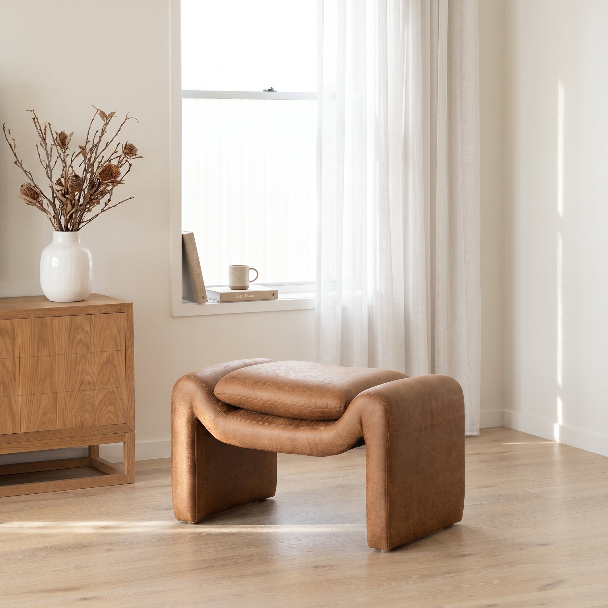 A modern low-profile ottoman stool upholstered in warm tan leather, positioned in a bright, minimalist room with light wood floors, a wooden sideboard, and a window with sheer curtains.
