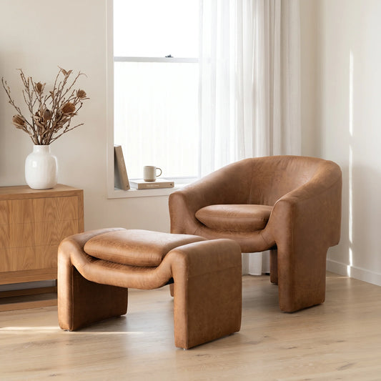 A modern curved armchair with a matching ottoman in a warm tan leather or faux leather, set in a bright, minimalist room with a wooden sideboard, window, and light-colored floor.