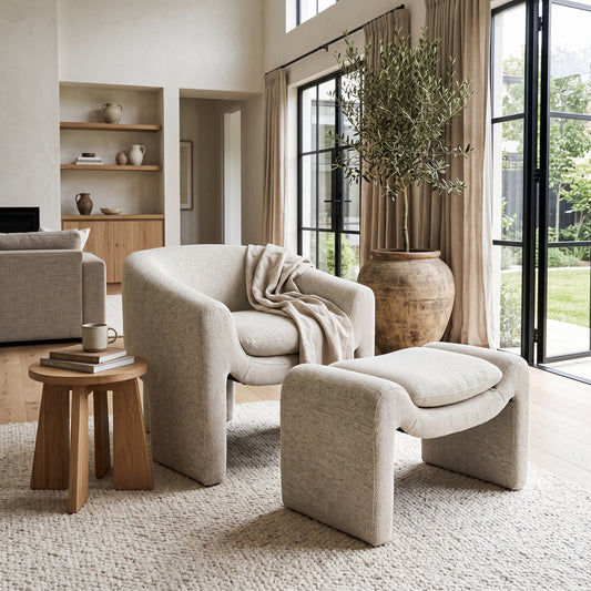 A modern upholstered lounge chair with a matching ottoman in a bright, neutral-toned living room. The chair and ottoman are upholstered in a light beige textured fabric, with a draped throw blanket on the chair.