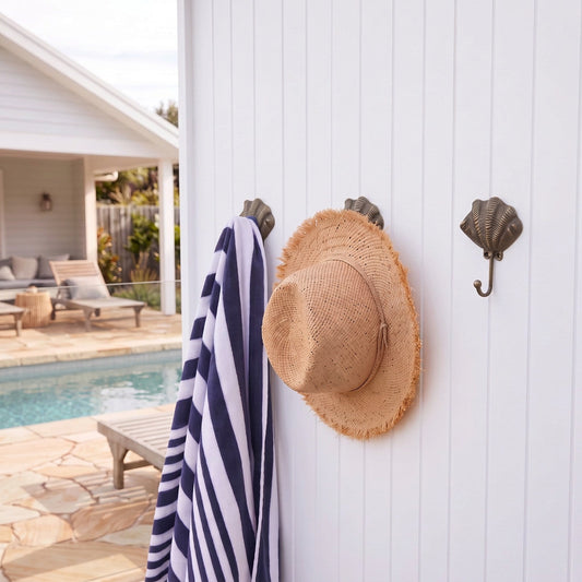 A woven straw sun hat and a navy and white striped beach towel hang from dark metal seashell wall hooks on a white vertical siding wall next to a swimming pool.