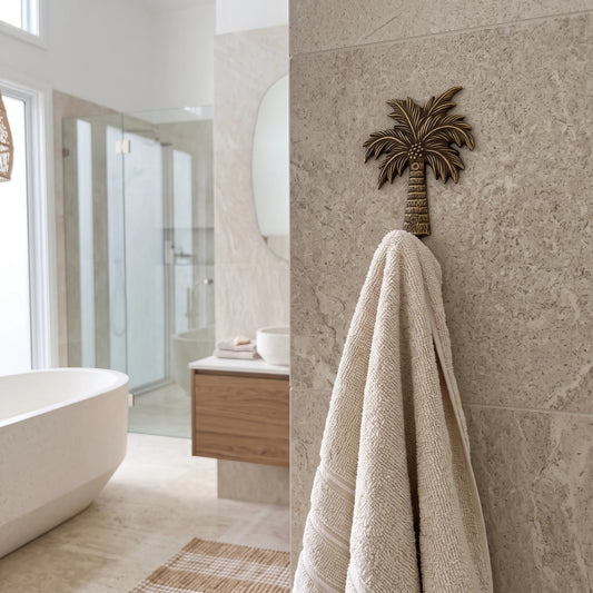 A textured cream towel hangs from a decorative bronze palm tree wall hook on a beige stone-tiled wall in a modern bathroom setting.
