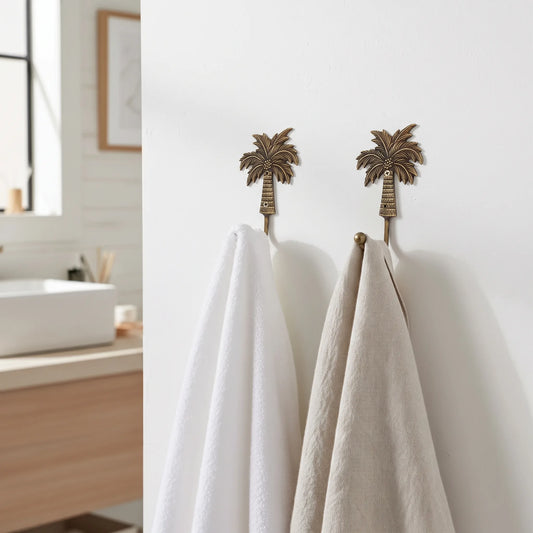 Two bronze palm tree wall hooks hanging on a white bathroom wall, holding a white towel and a light beige towel.