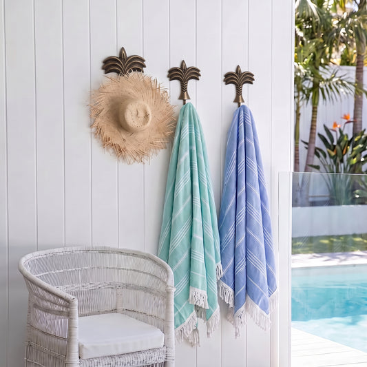 Two woven beach towels, one mint green and one light blue, hanging from dark bronze palm tree wall hooks on a white vertical wood paneled wall. A straw hat rests on the left hook, and a white wicker chair is visible in the foreground.