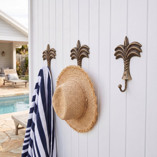 Three bronze palm tree wall hooks mounted on a white shiplap wall, with a navy and white striped towel and a straw hat hanging from them.