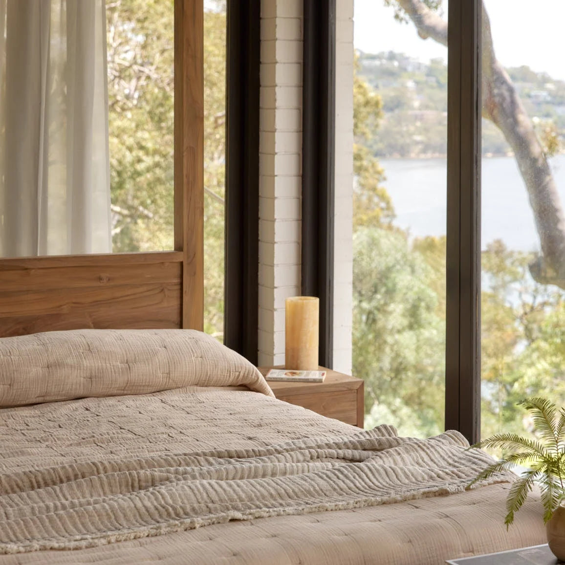 A neatly made bed with textured beige bedding and a wooden headboard, positioned in a bright bedroom with large windows overlooking a scenic lake and forest view.